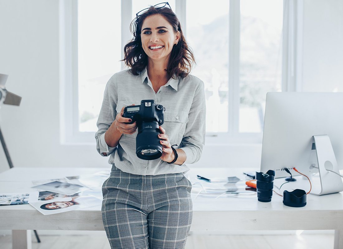 Insurance by Industry - Happy Photographer Holds Her Camera as She Leans on Her Desk at Her Office